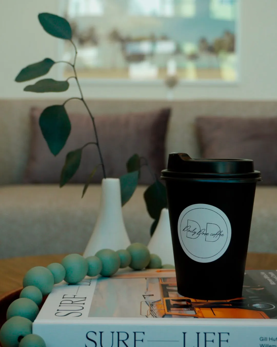 A black Daily Dose cup with its round logo sticker, resting on a Surf Life book beside a eucalyptus stem in the venue lounge
