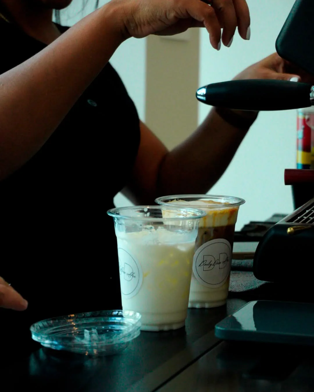 A barista works the espresso machine behind two clear cups sealed with round Daily Dose stickers, one a pale blended drink and one an iced caramel latte